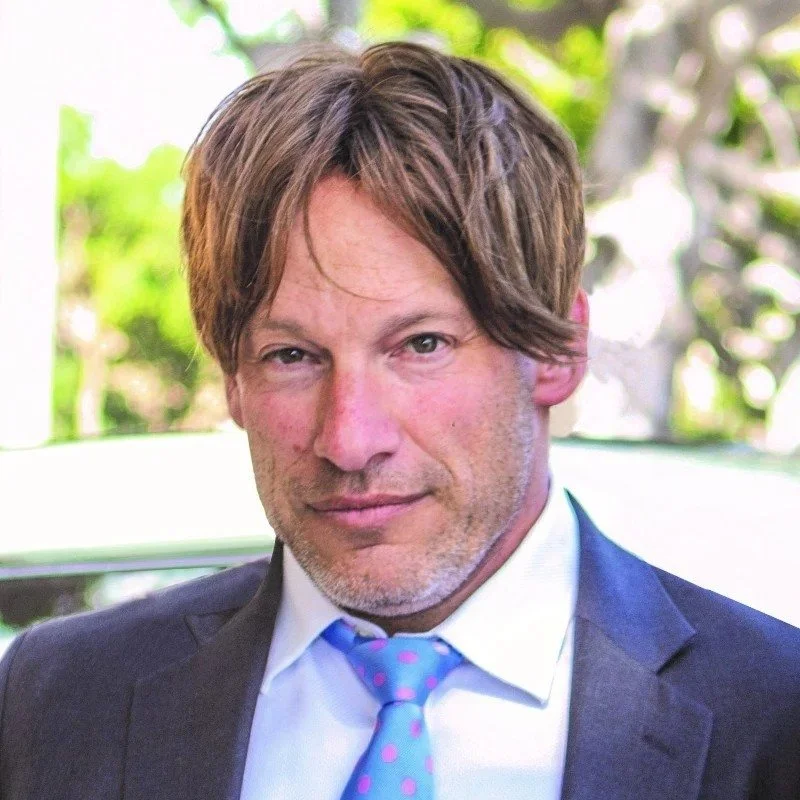 A man with medium-length brown hair, wearing a white shirt, blue tie with pink polka dots, and a dark suit jacket, standing outdoors with blurred greenery in the background.