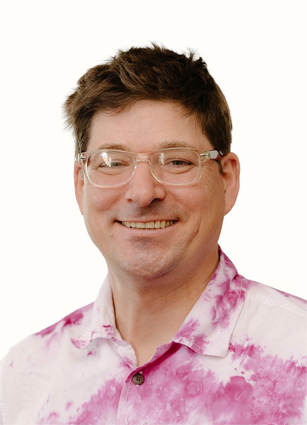 A man with short brown hair, wearing clear glasses and a pink and white tie-dye shirt, smiling at the camera.