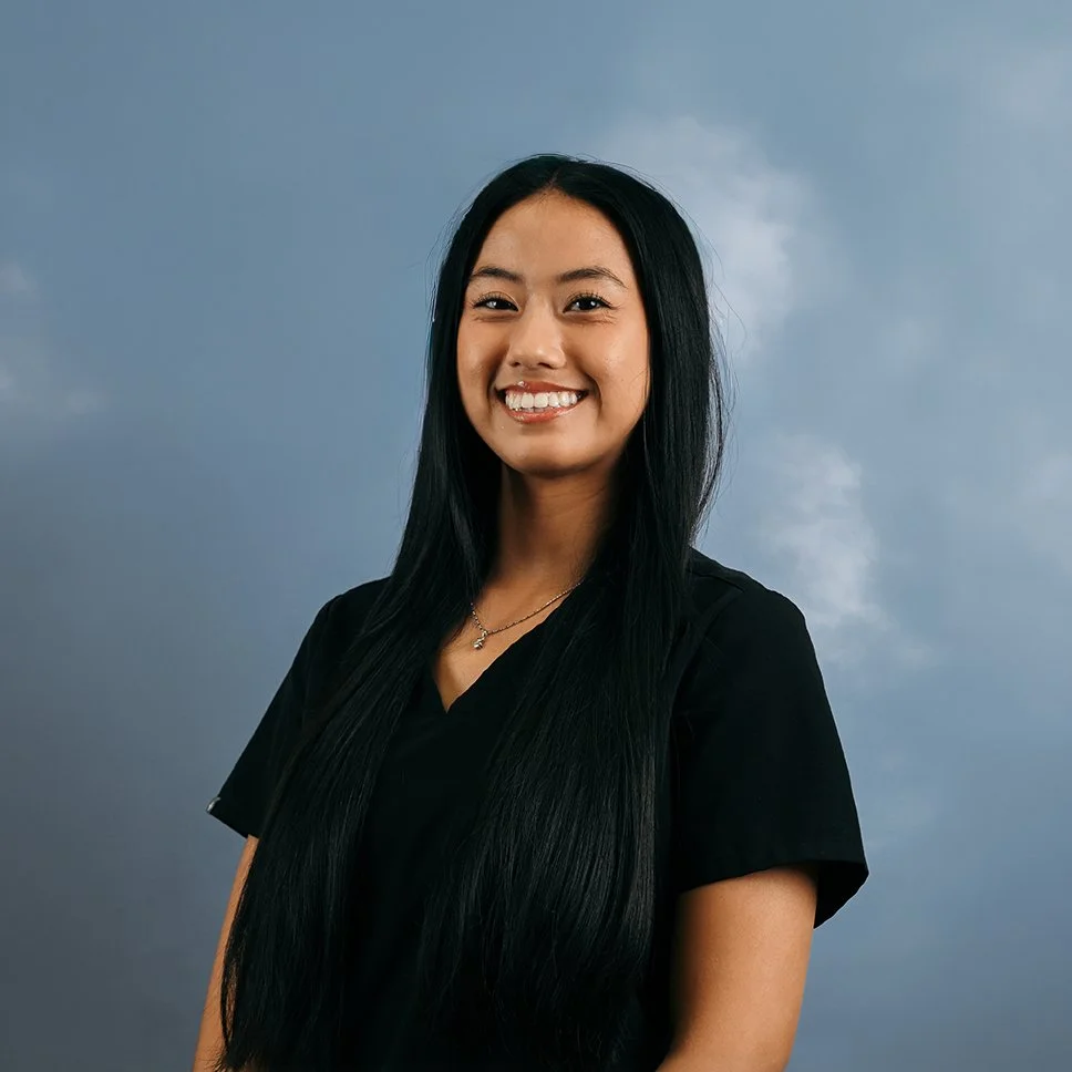Young woman with long black hair smiling, wearing a black shirt, against a cloudy sky background.