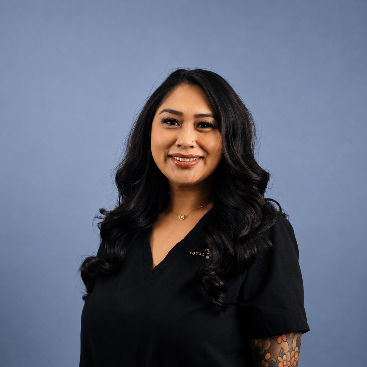 A woman with long black wavy hair smiling against a light blue background. She is wearing a black top and has a tattoo sleeve on her right arm.