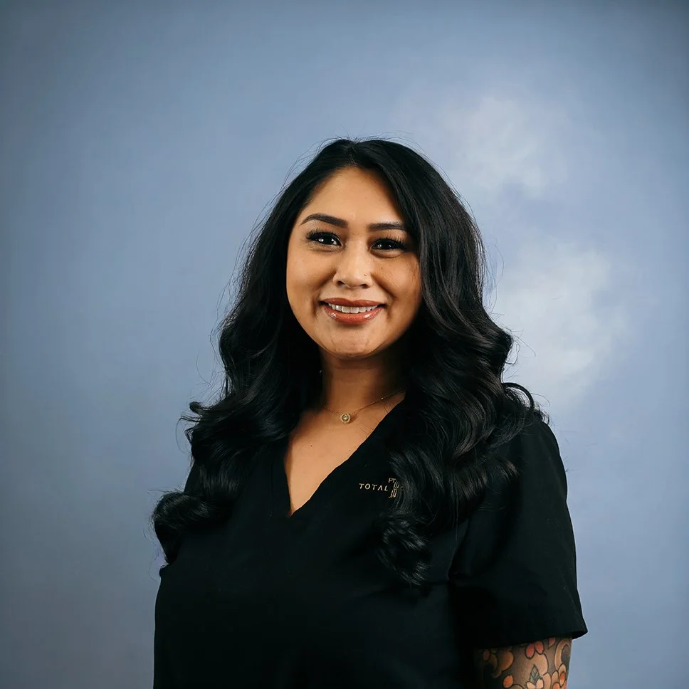 A woman with long black wavy hair smiling against a light blue background. She is wearing a black top and has a tattoo sleeve on her right arm.