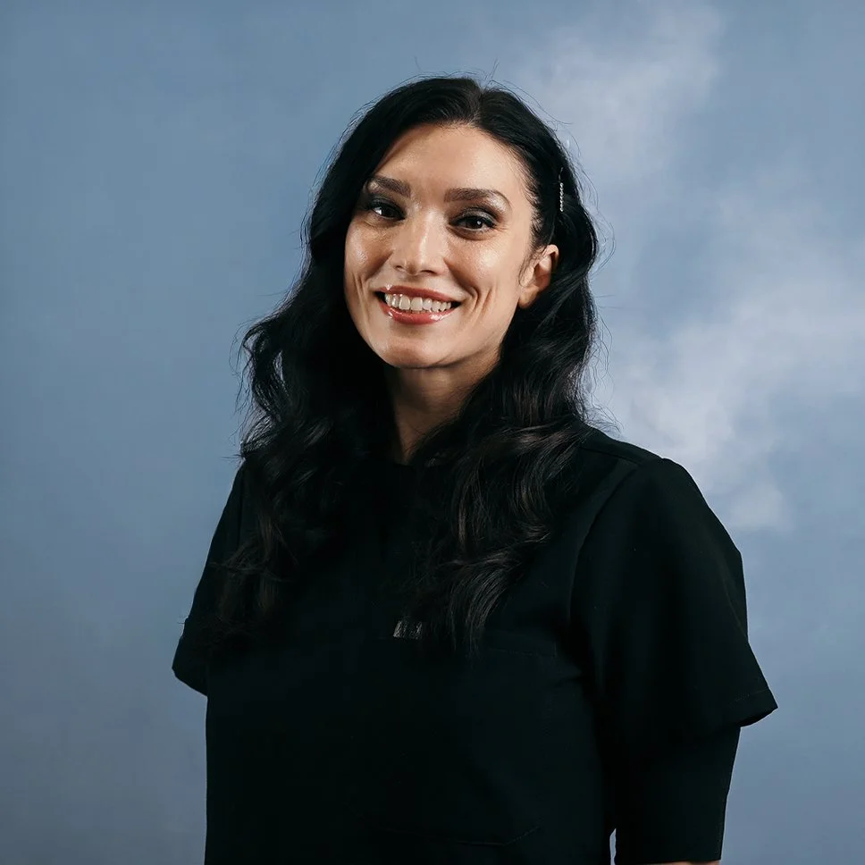 A woman with long black wavy hair, smiling, wearing a black top, against a cloudy blue sky background.