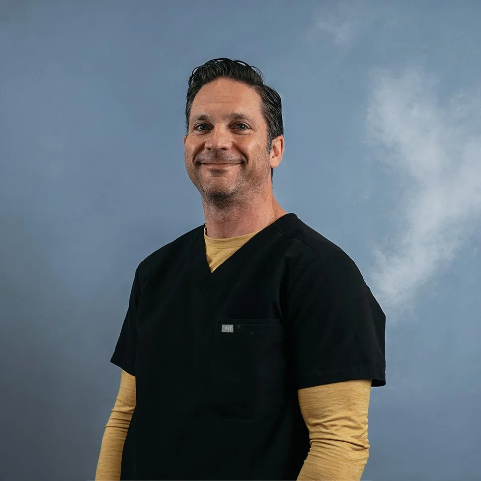 A middle-aged man with dark hair, wearing black scrubs and a beige long-sleeve shirt underneath, standing against a blue-gray background.