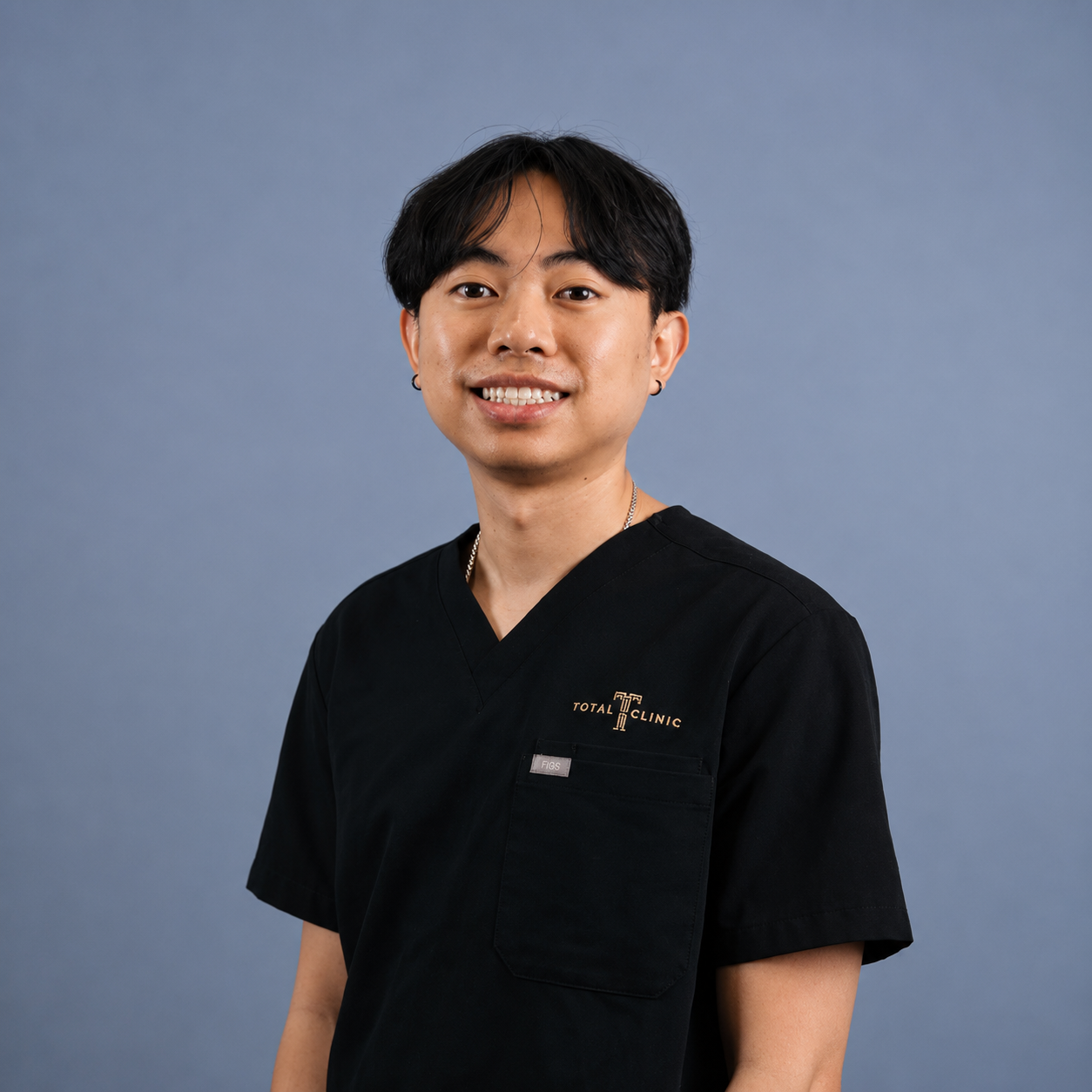 A man smiling and wearing a black Medical Clinic uniform against a blue cloudy background.