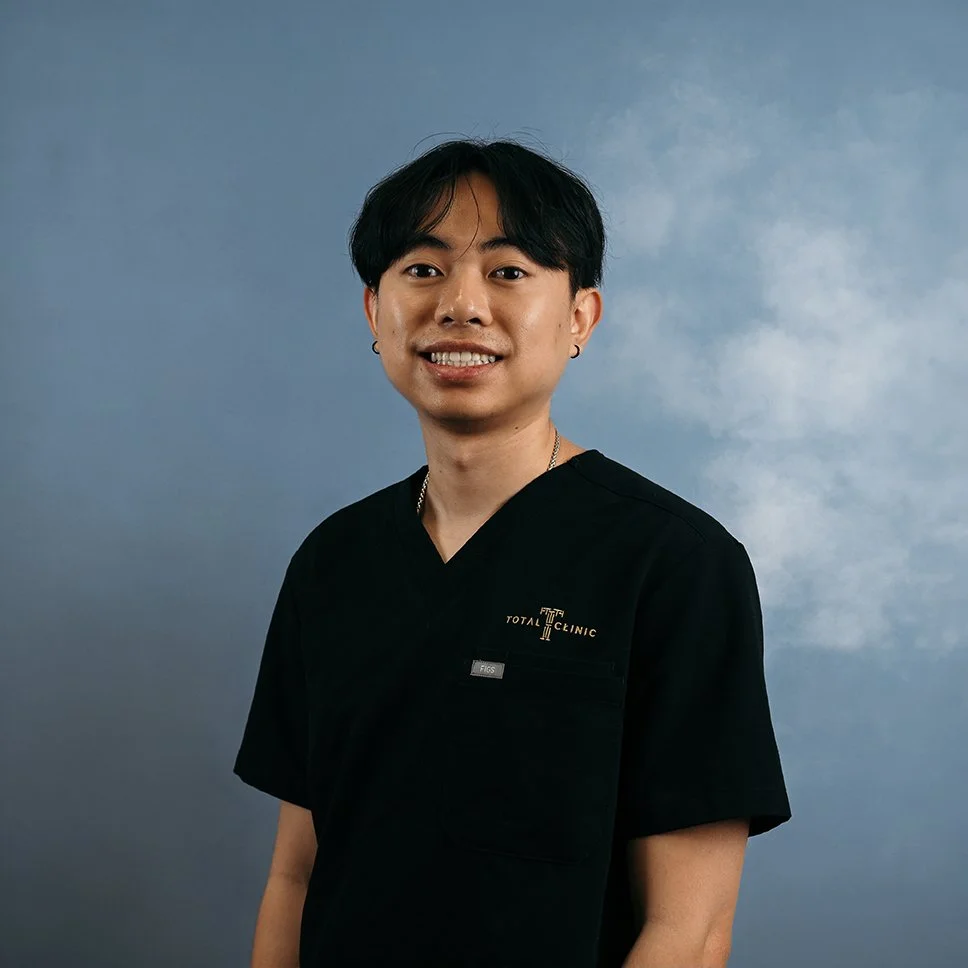 A man smiling and wearing a black Medical Clinic uniform against a blue cloudy background.