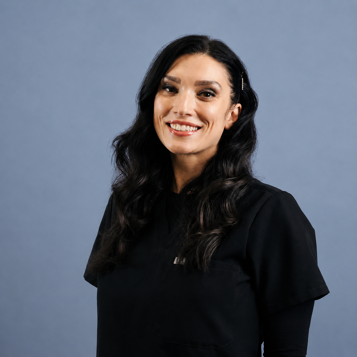 A woman with long dark wavy hair smiling, wearing a black top, standing against a plain blue background.