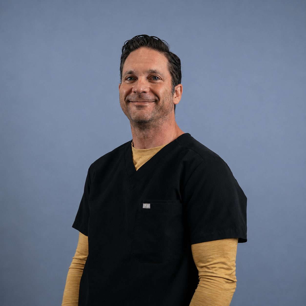 A man wearing black medical scrubs with a tan long sleeve shirt underneath. He is smiling and standing against a plain blue background.