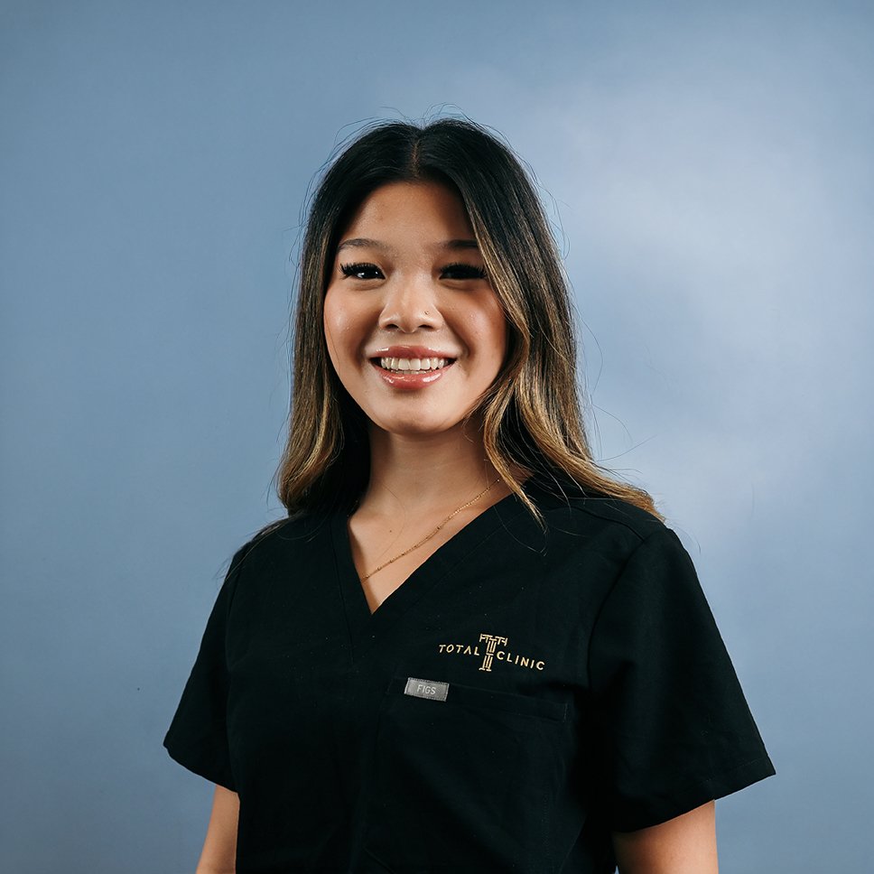 A smiling woman with shoulder-length dark hair and a light brown streak, wearing a black medical scrubs top with the logo 'TOTAL CLINIC' and 'FIGS' on it, standing against a blue background.