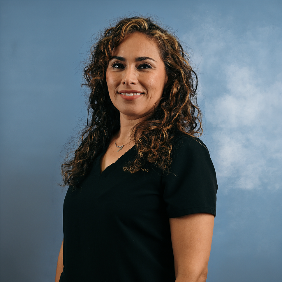 A woman with curly brown hair wearing a black medical scrubs uniform, smiling against a blue background.