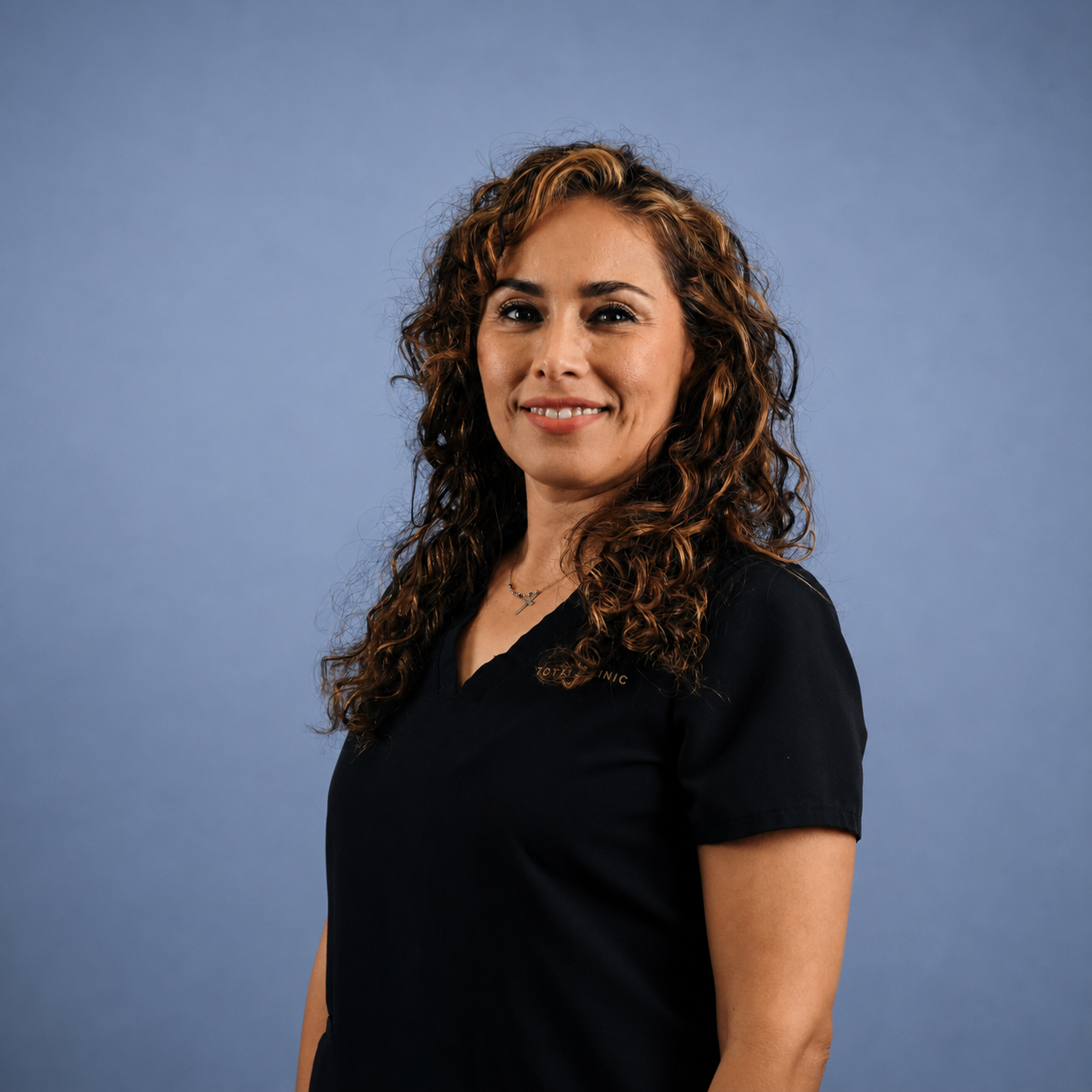A woman with curly brown hair wearing a black medical scrubs uniform, smiling against a blue background.