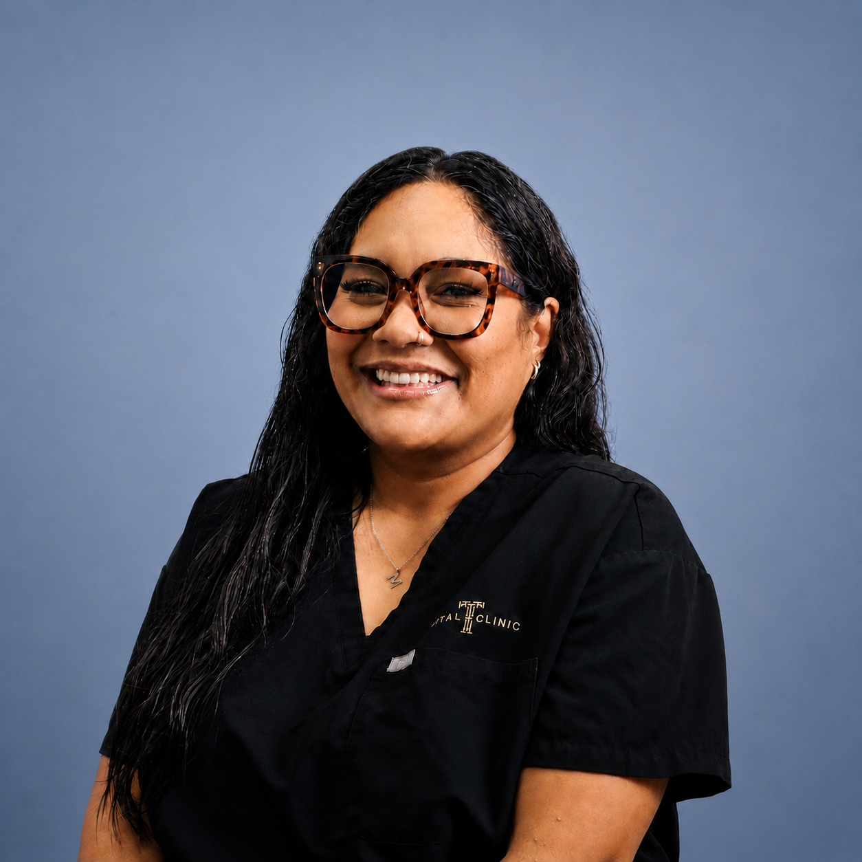 A woman with long black hair, wearing glasses, a nose ring, a necklace, and a black uniform with a logo that says 'Dental Clinic,' smiling against a blue background.
