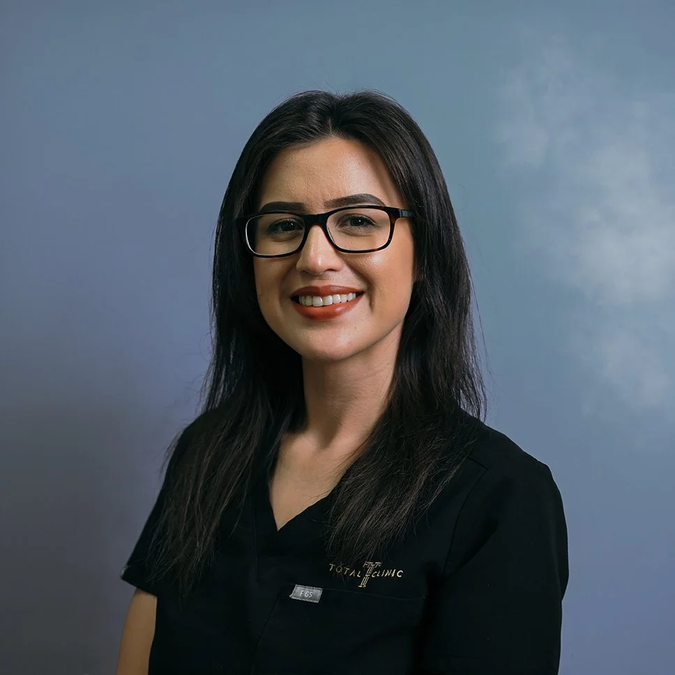 A woman with long dark hair, glasses, and a black uniform smiling at the camera against a blue background.
