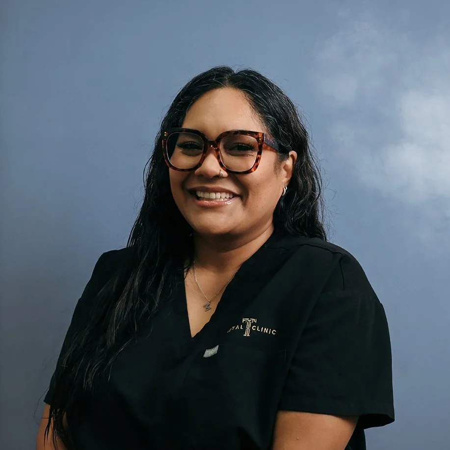 A woman with long black hair, wearing glasses, a nose ring, a necklace, and a black uniform with a logo that says 'Dental Clinic,' smiling against a blue background.