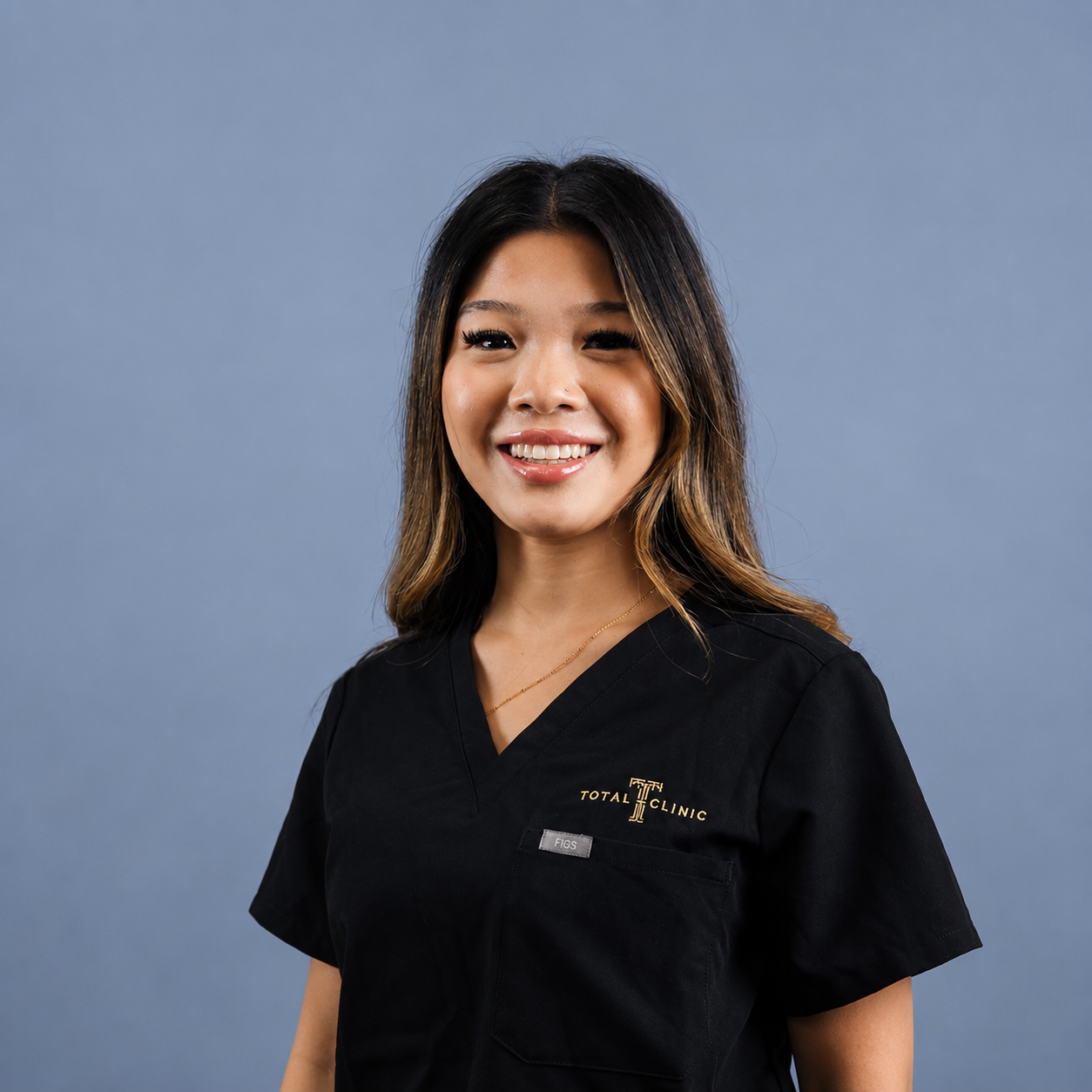 A smiling woman with shoulder-length dark hair and a light brown streak, wearing a black medical scrubs top with the logo 'TOTAL CLINIC' and 'FIGS' on it, standing against a blue background.