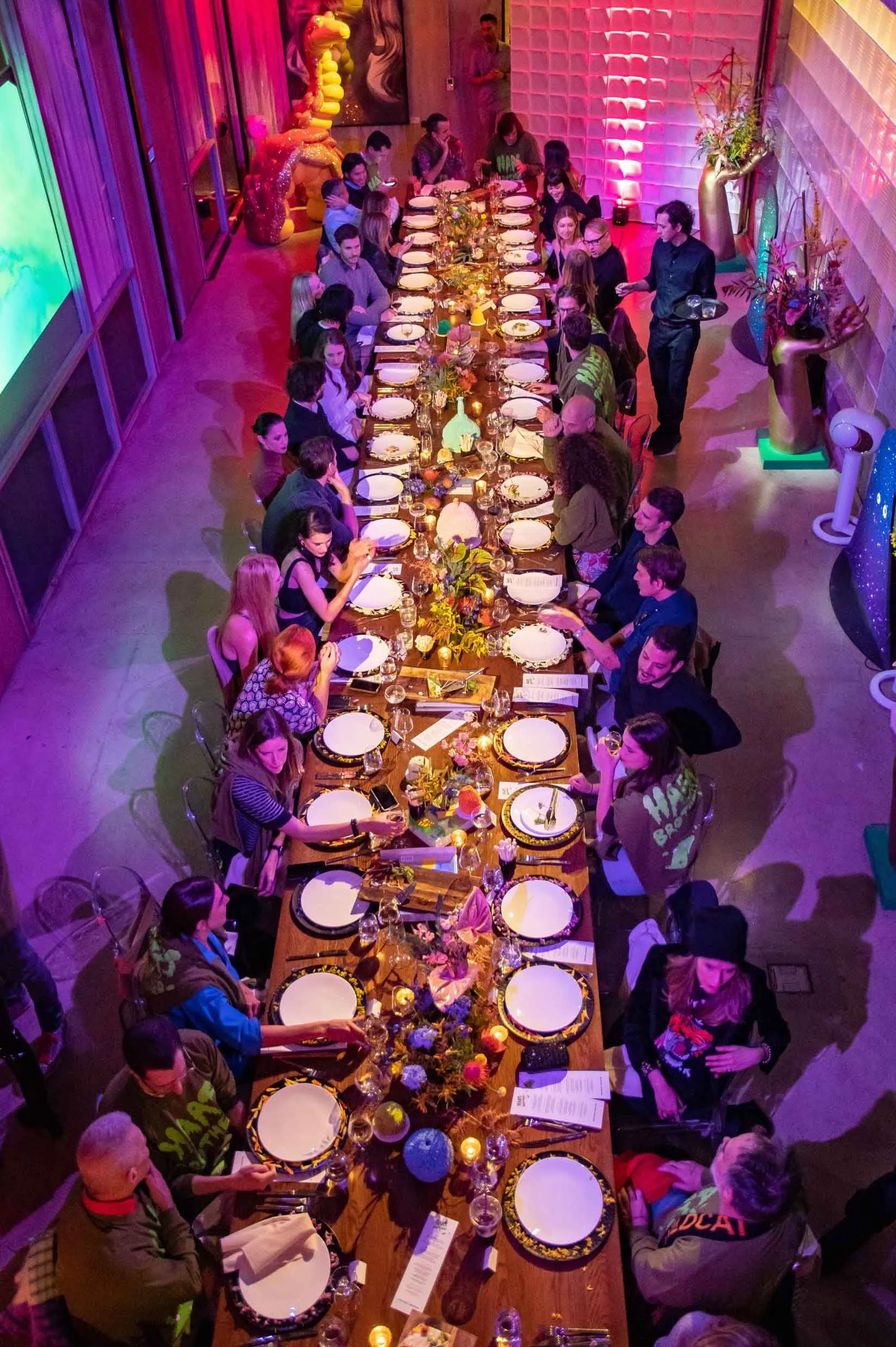 A long, decorated dining table set for a large gathering with plates, glasses, and floral centerpieces, in a colorful, decorated room with people seated and standing around.