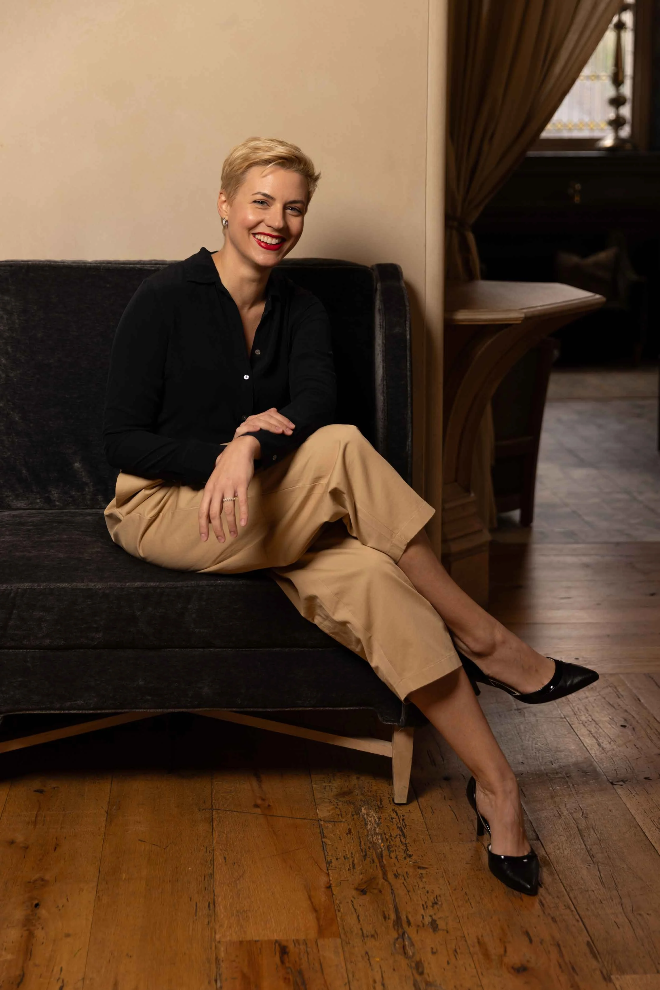 A woman sitting on a black sofa in a room with wooden floors and curtains, smiling and wearing a black blouse, beige pants, and black high heels.