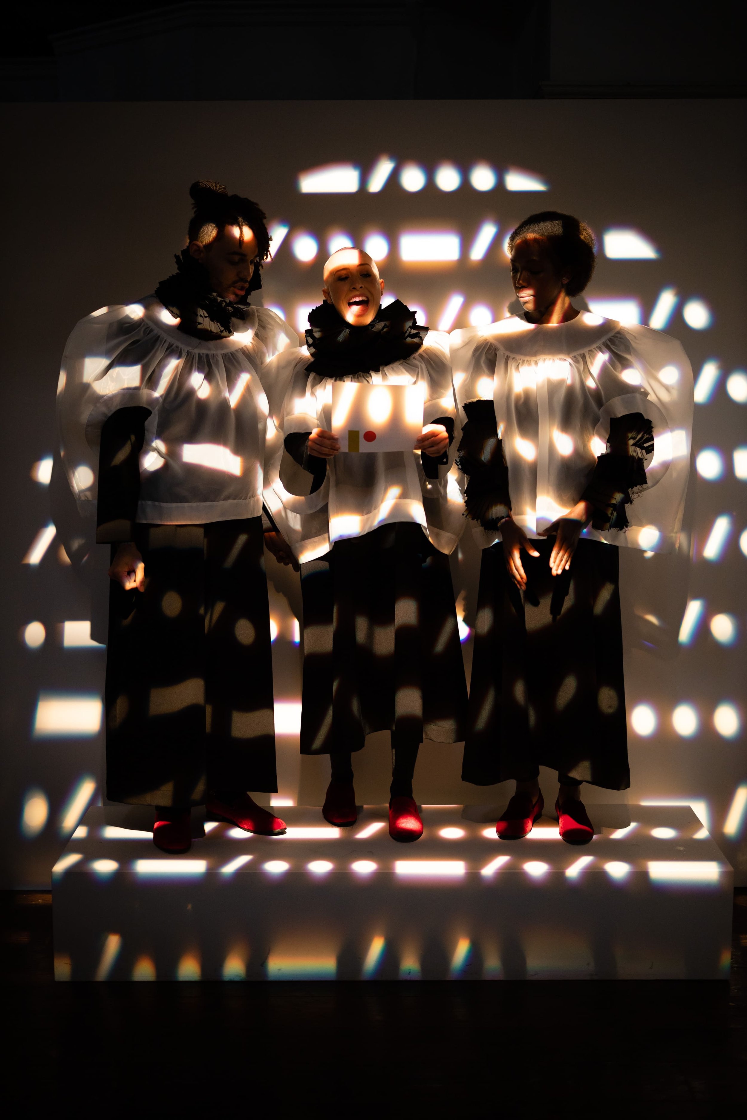 Three women dressed in white blouses with black ruffled collars, black skirts, and red shoes, standing on a platform with light reflections and shadows projected on their faces and clothing.