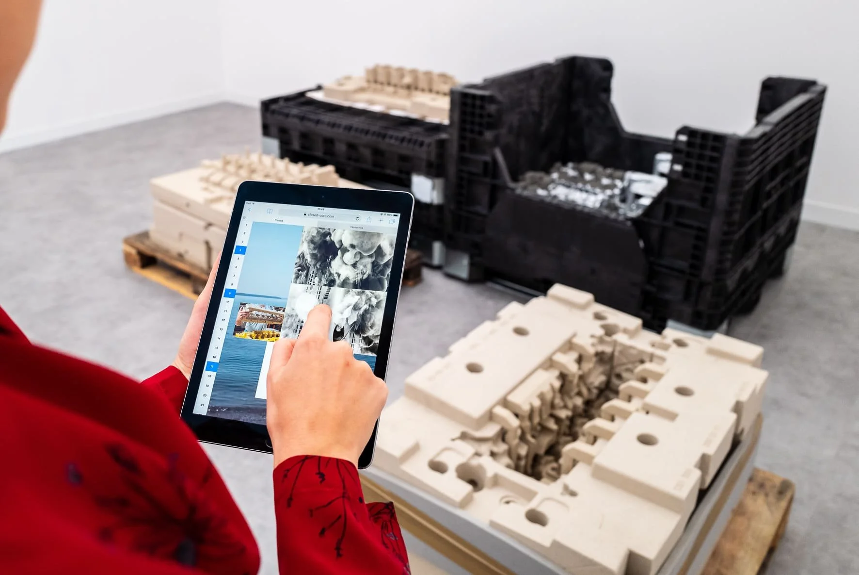 Person in a red shirt using a tablet to browse images in an art installation featuring architectural models and abstract sculptures.