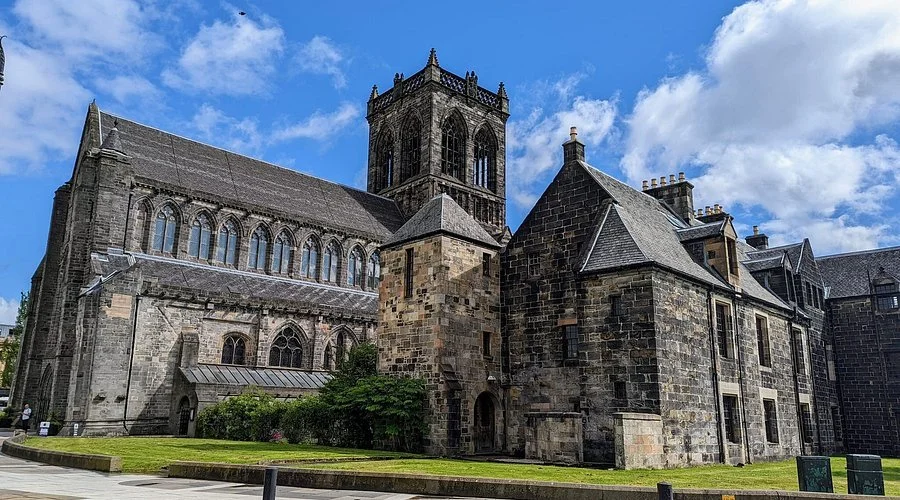 A historic stone church with gothic architecture, tall tower, and surrounding stone buildings against a blue sky with clouds.