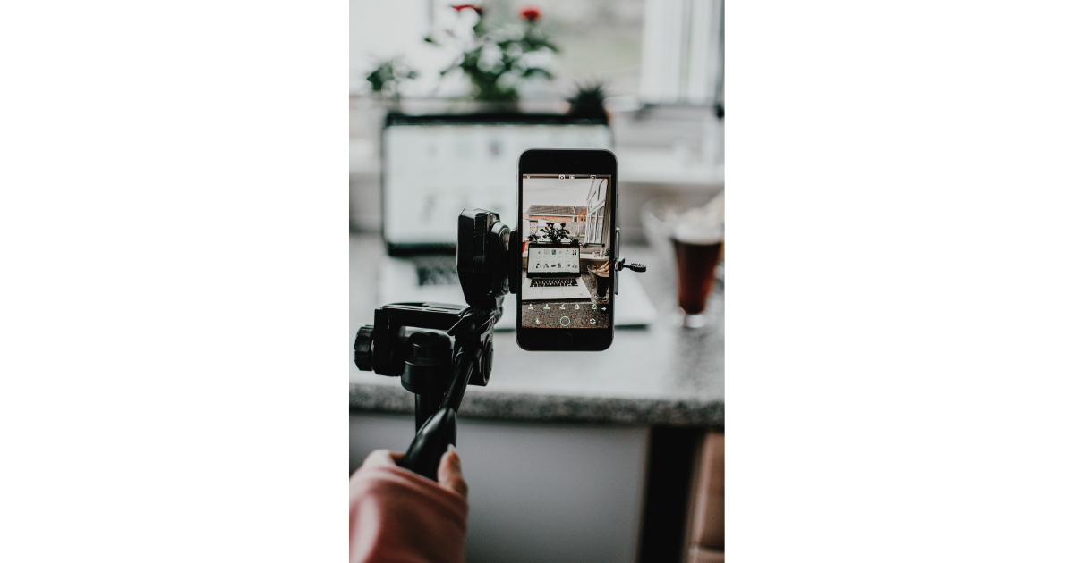 Person is filming a setup of a laptop, a small plant, and a glass of iced tea on a kitchen counter using a camera mounted on a selfie stick.