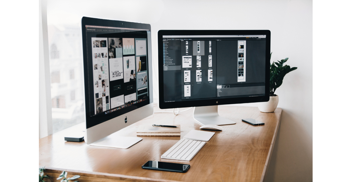 Two Apple desktop computers on a wooden desk, one displaying a grid of images and the other displaying design files, with a keyboard, mouse, smartphone, notebook, pen, and a potted plant nearby.