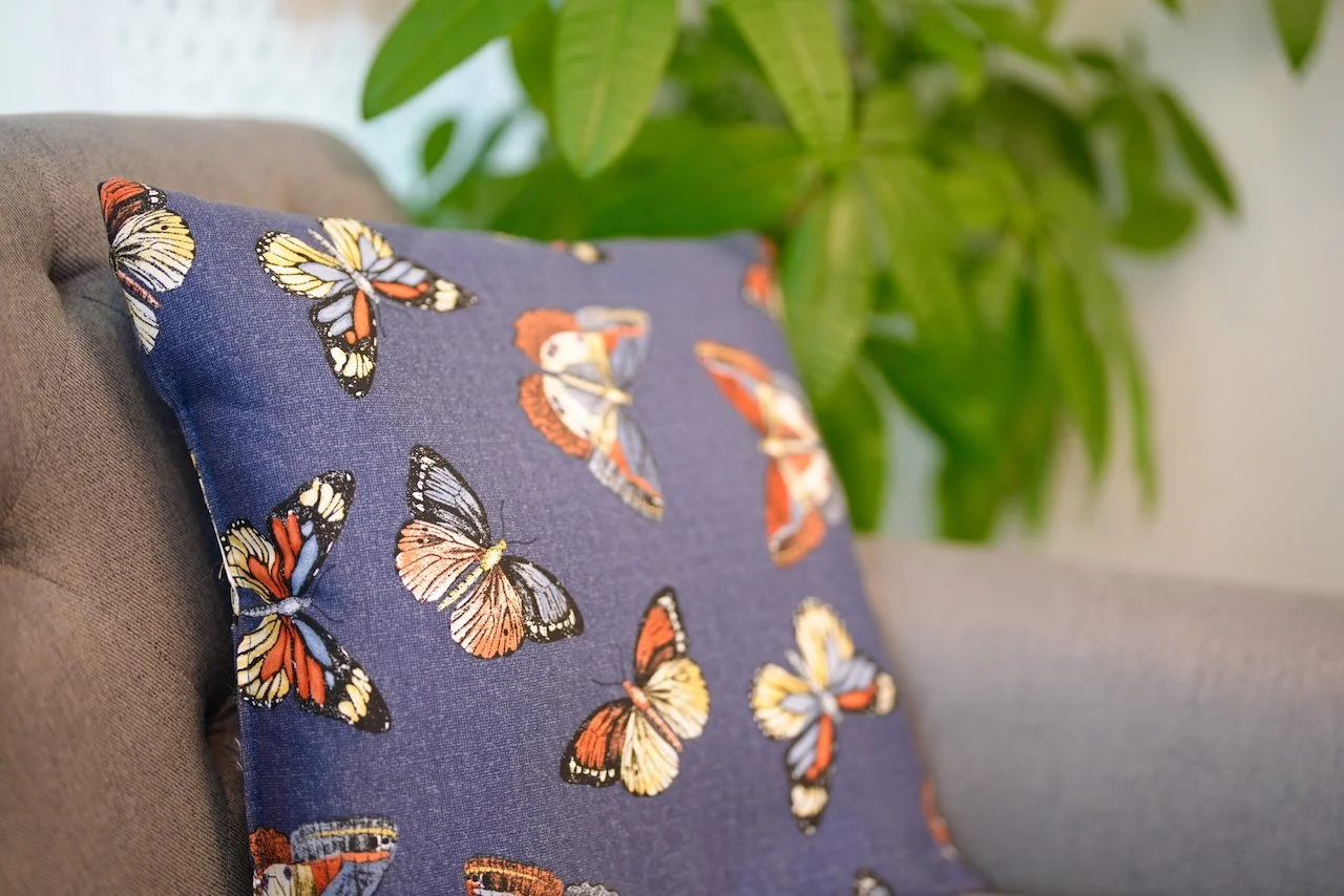 A decorative pillow with butterfly patterns on a sofa, with a large green plant in the background.