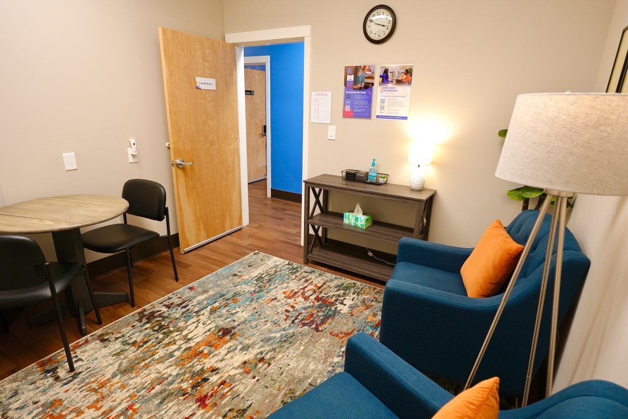 A cozy waiting room with two blue armchairs with orange pillows, a wooden side table with a lamp and sanitizer, a small round table with black chairs, colorful patterned rug, a wall clock, and informational posters.