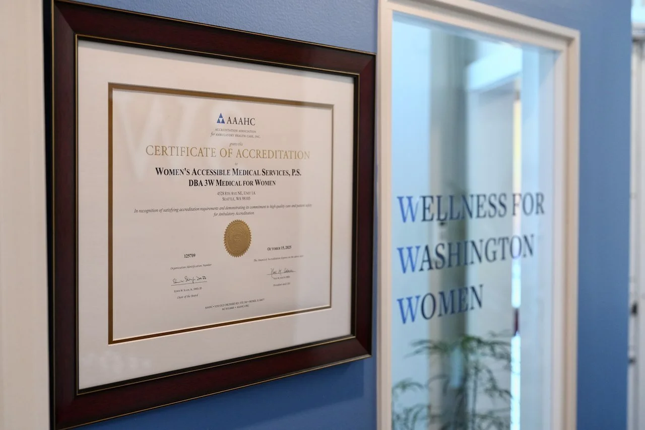 A framed certificate of accreditation for Women's Accessible Medical Services, displayed on a blue wall next to a glass door with blue lettering that reads Wellness for Washington Women.