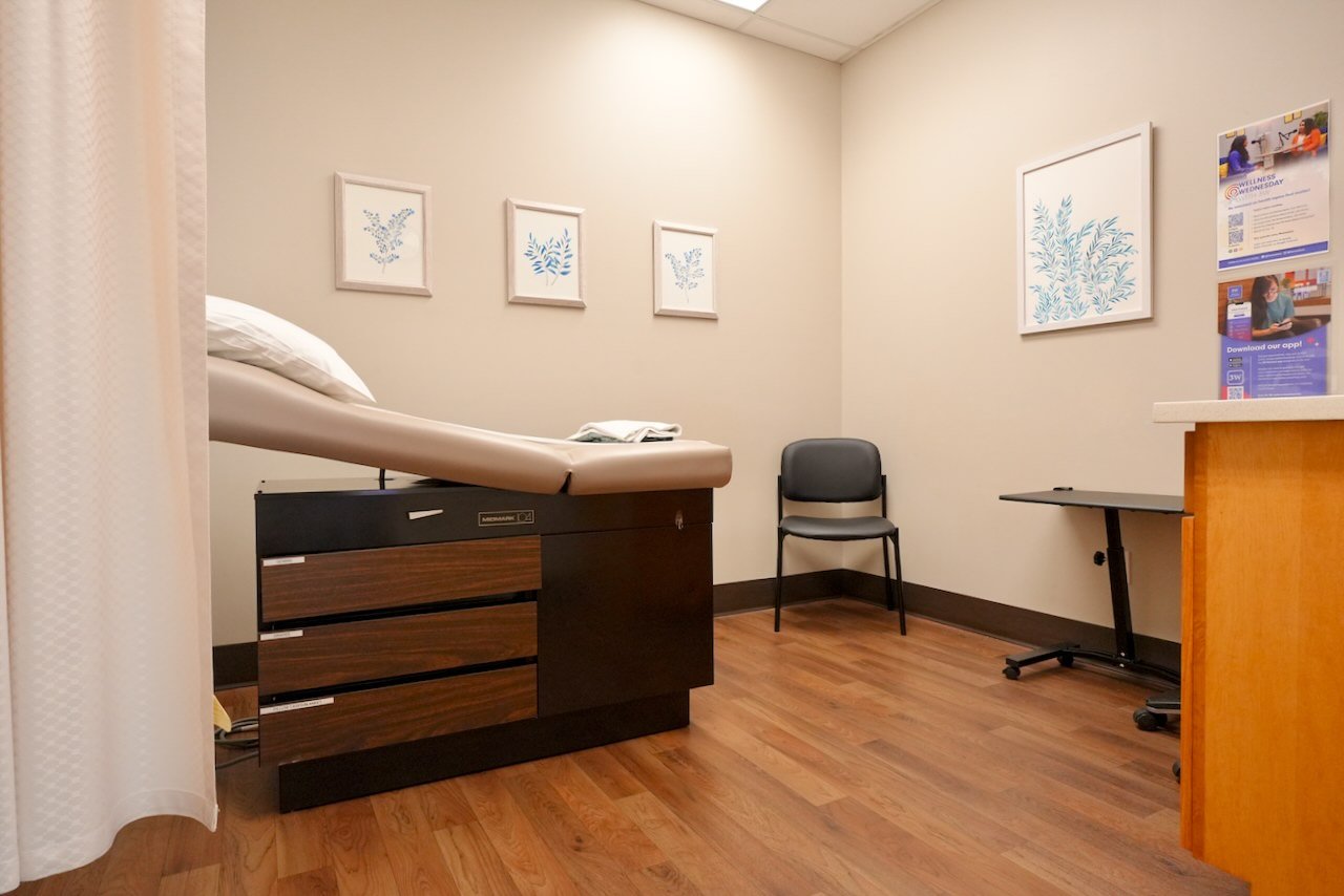 A medical examination room with an exam bed, a black chair, a small table, and framed botanical artwork on the walls.