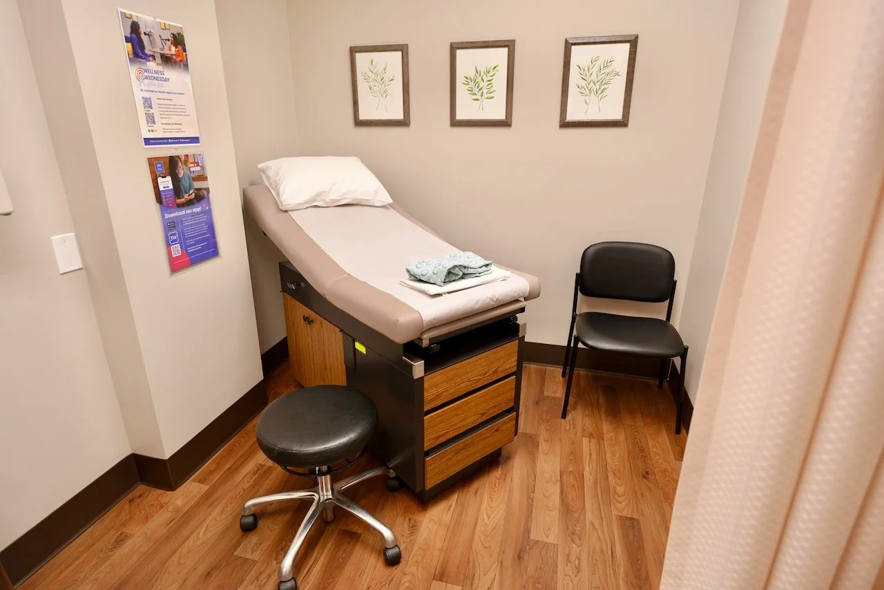 A medical examination room with an exam table covered with a pillow and folded towels, two chairs, and framed botanical prints on the wall.