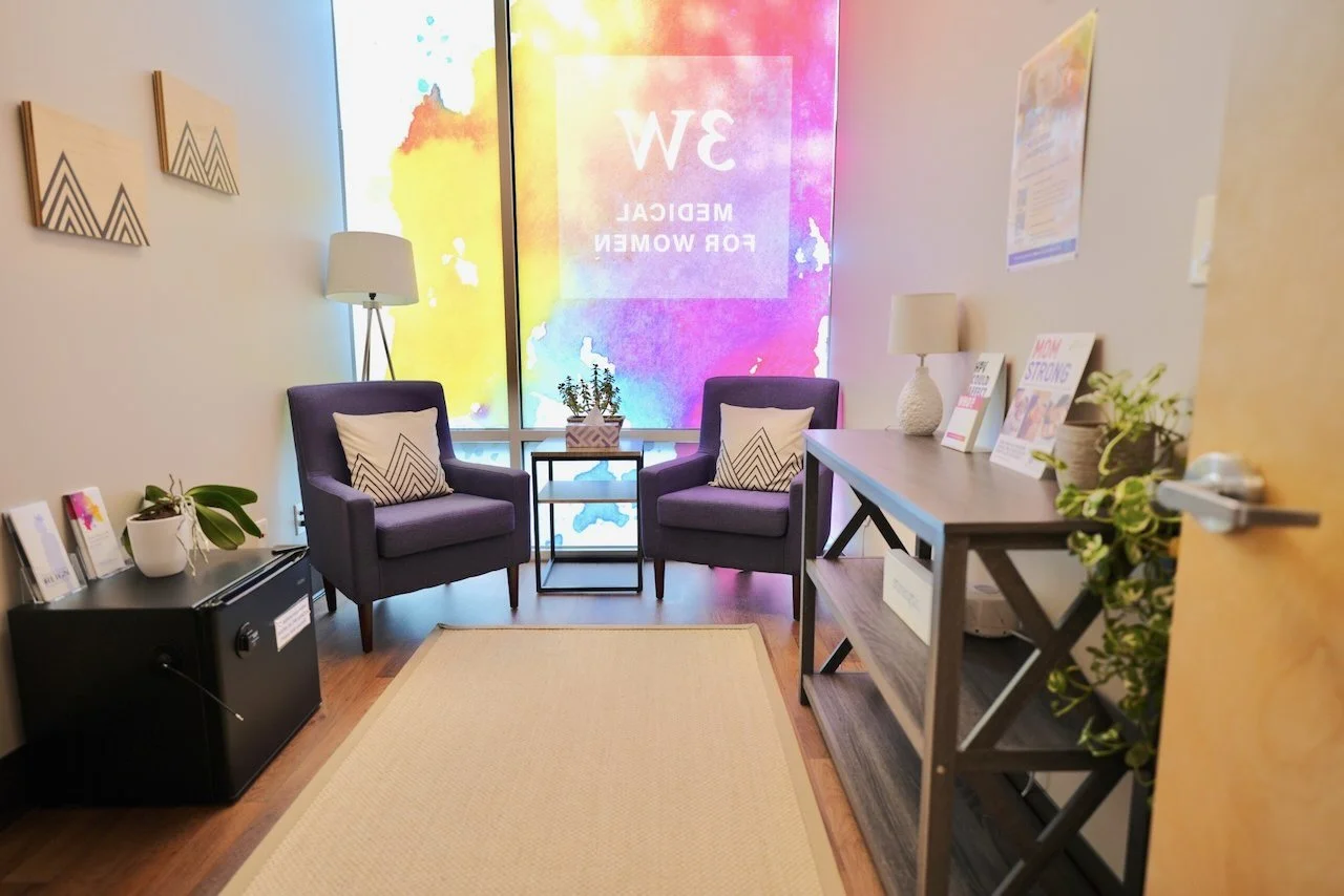 A cozy waiting room with two purple armchairs, each with a decorative pillow, a small table with a plant between them, a black cabinet with potted plants and magazines, a wooden console with magazines and a lamp, and a large, colorful digital screen 