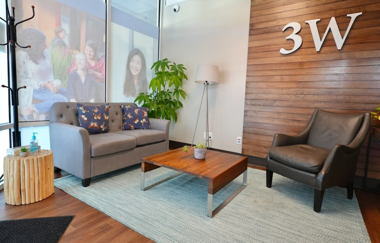 Modern office reception area with a gray sofa with butterfly-printed pillows, a black leather armchair, a wooden coffee table with a small potted plant, a tall floor lamp, a green plant, a patterned rug, a window with photos, a wooden accent wall wit