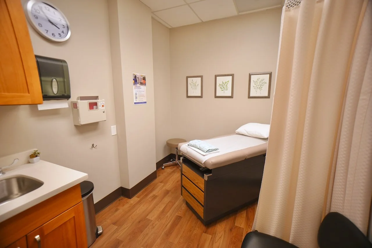 Hospital examination room with a bed, framed artwork of botanicals on the wall, bedside stool, small kitchenette with a sink, wall clock, and privacy curtains.