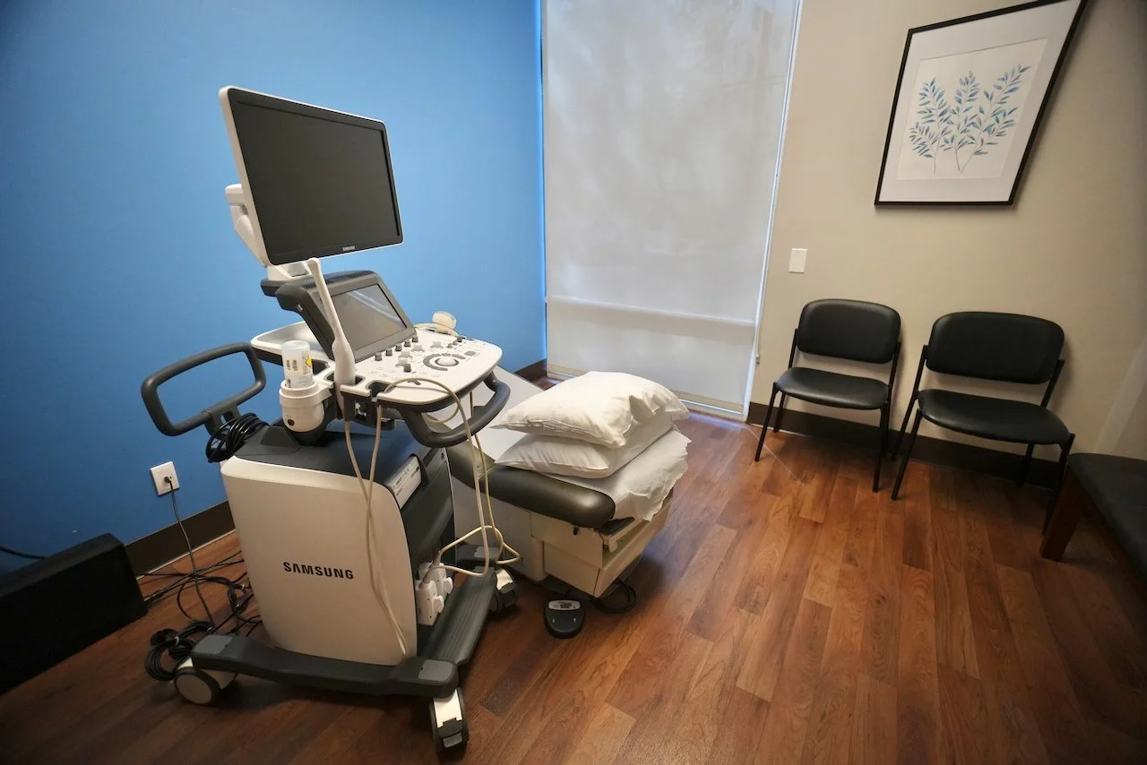 Medical examination room with an ultrasound machine, two pillows on an examination table, and three chairs along the wall.