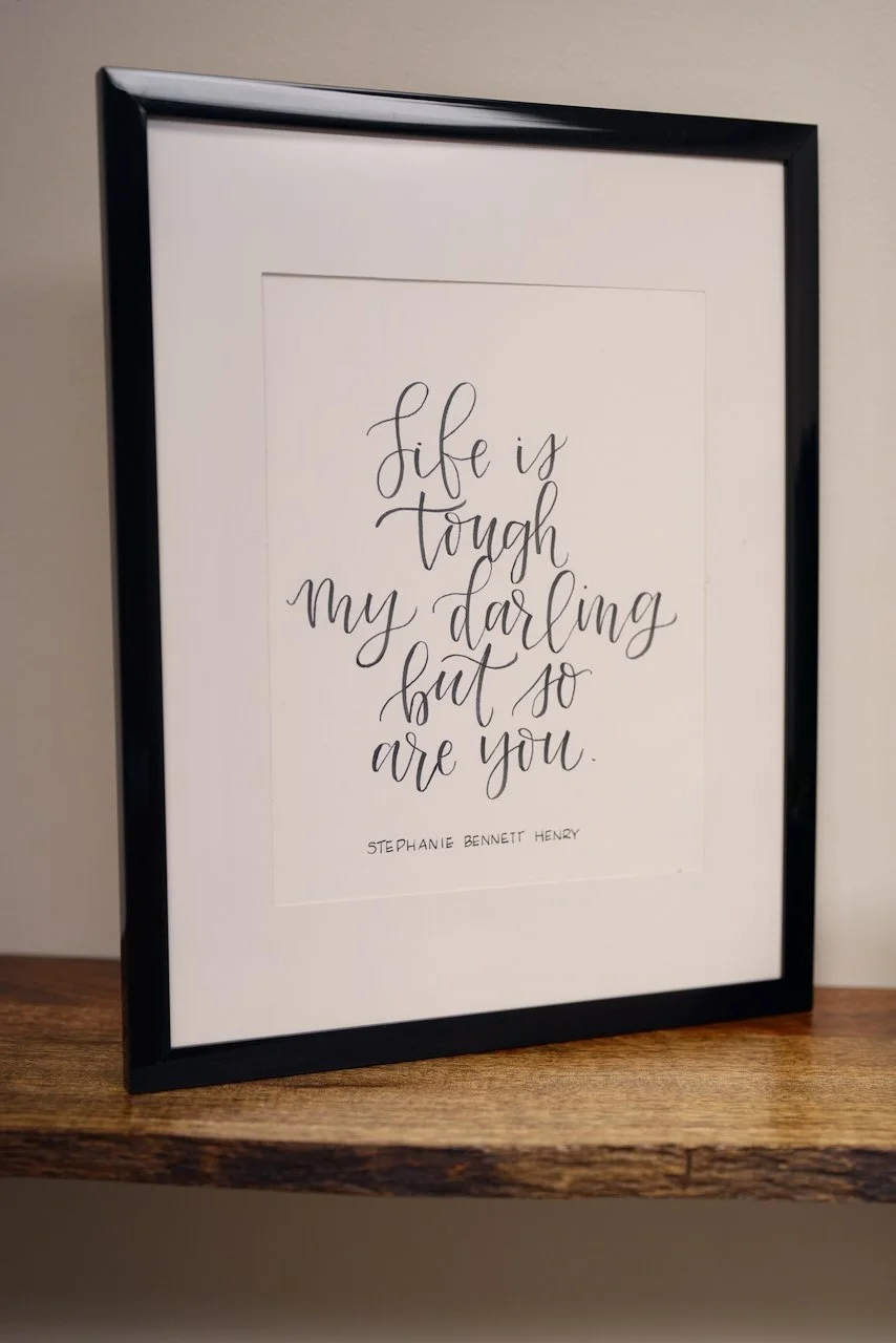 Framed print with the quote "Life is tough my darling but so are you" by Stephanie Bennett Henry placed on a wooden shelf.