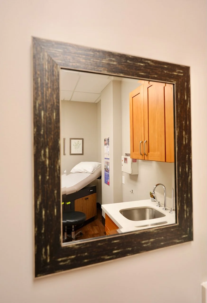 Reflection of a hospital room in a mirror showing a bed, wooden cabinets, and a small sink.