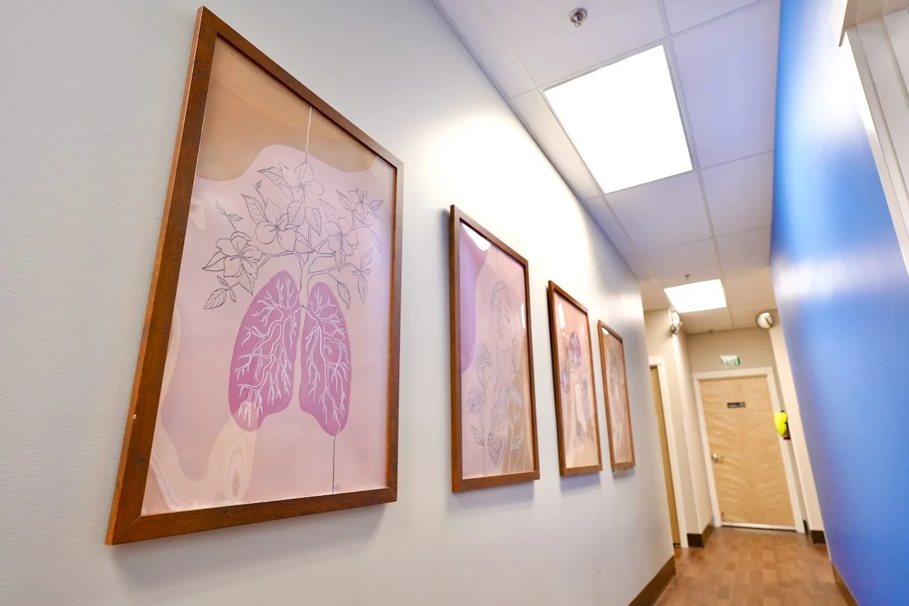A hallway with several framed botanical illustrations hanging on the wall. The first frame features a pink outline of a plant with roots and leaves, and the others contain similar botanical artwork. The hallway has a white ceiling with fluorescent li