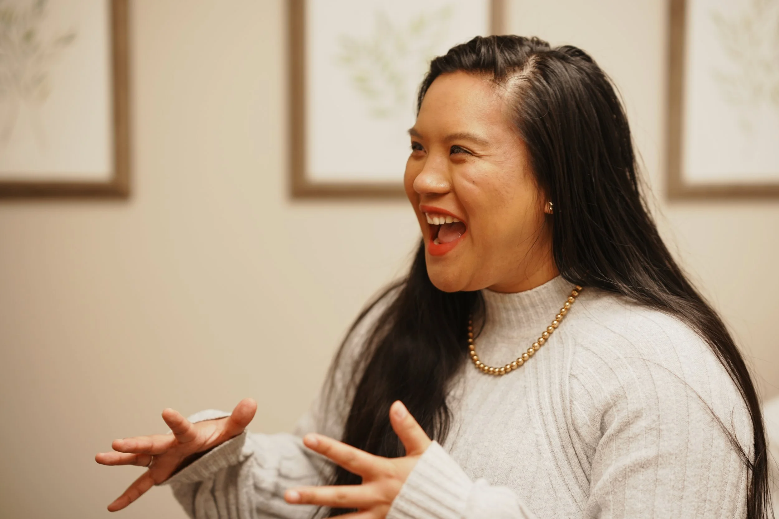 A woman with long dark hair laughing and gesturing with her hands, indoors with framed artwork on the wall behind her.