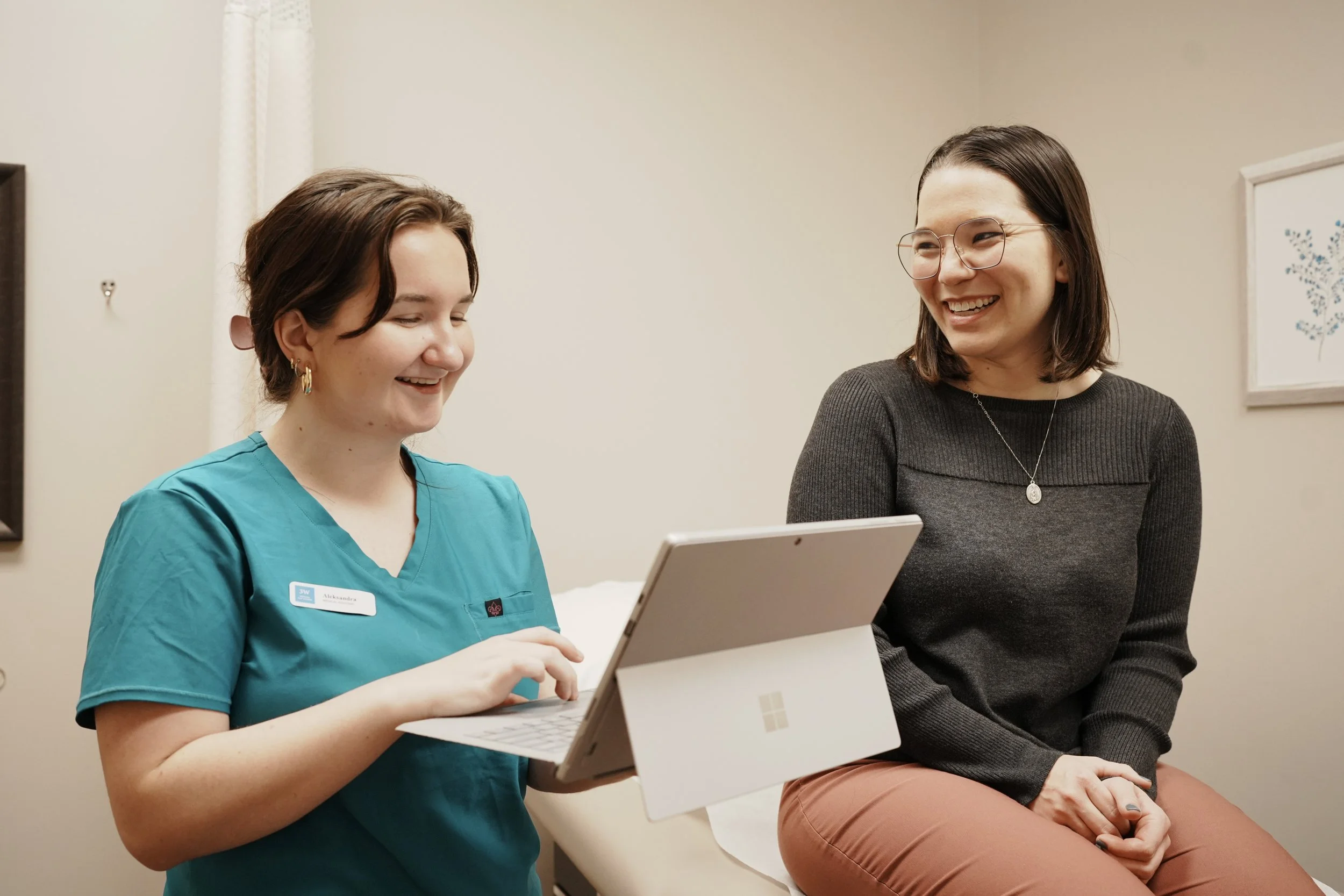 A nurse showing a tablet to a woman in a medical examination room, both smiling and engaging in conversation.
