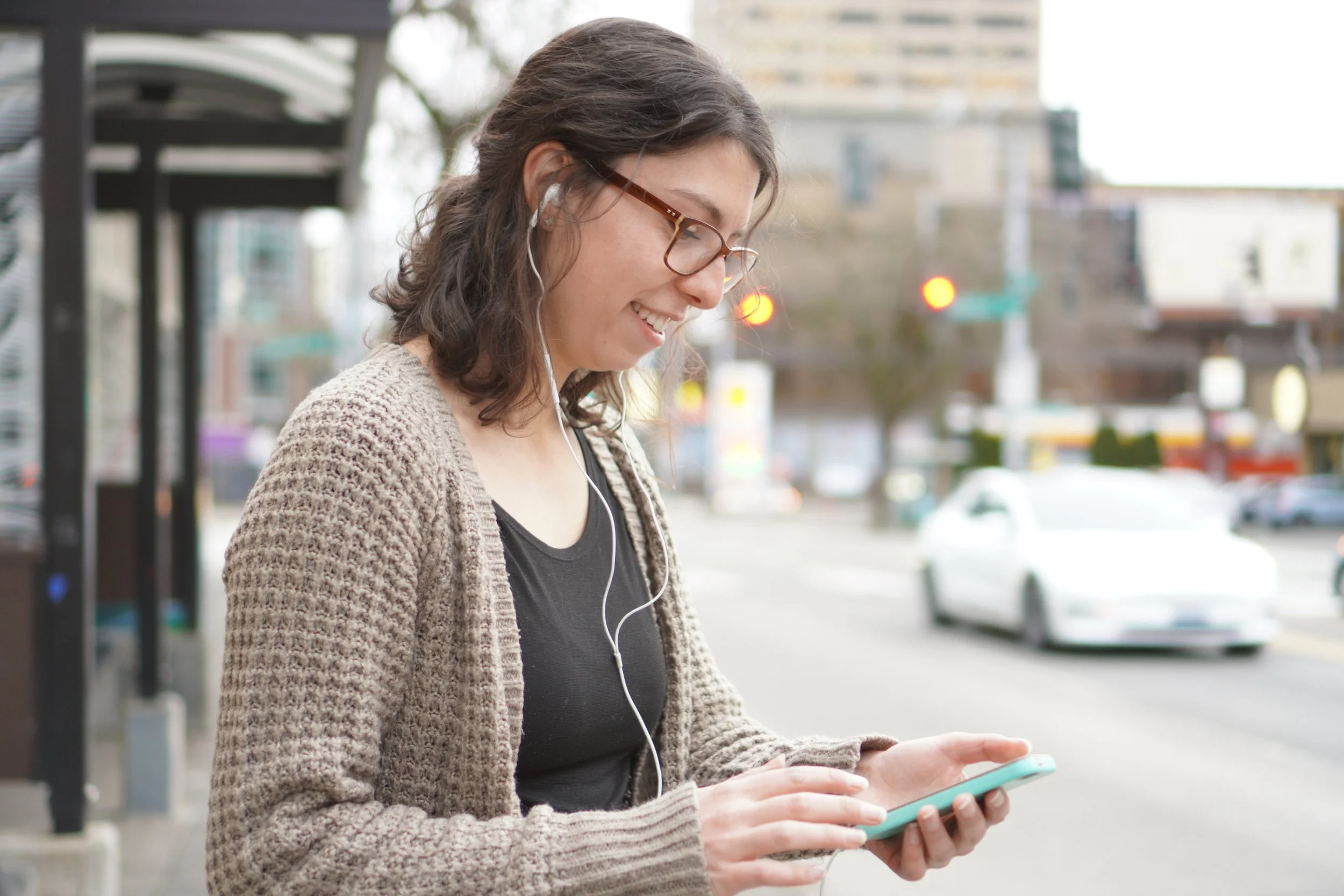 Young woman with glasses and earphones, using a smartphone outdoors on a city street.