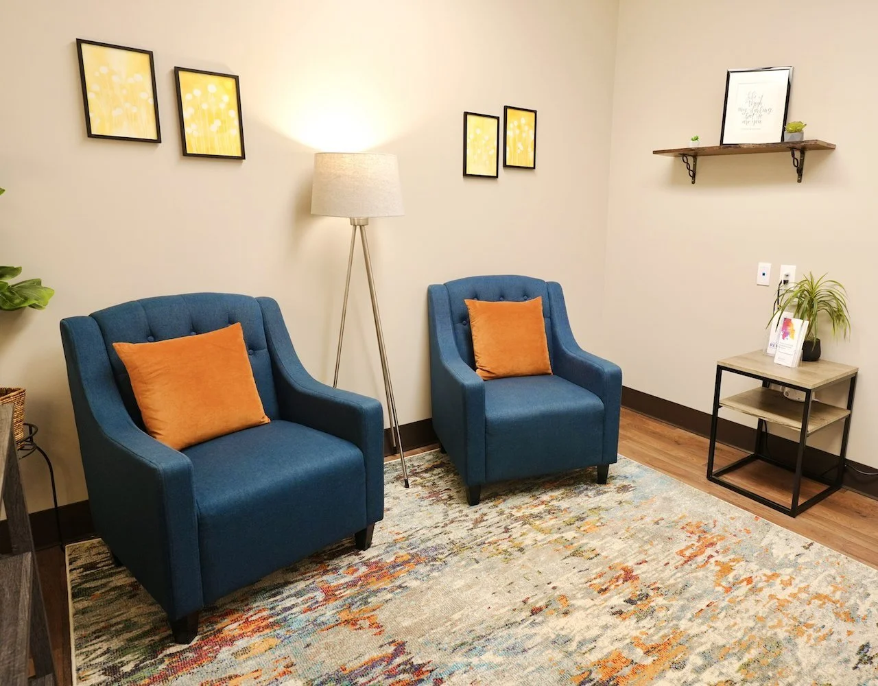 A cozy waiting room with two blue armchairs each with an orange cushion, a tall floor lamp, a small wooden side table with a plant and brochures, a floating shelf with decor, framed artwork, and a patterned area rug.
