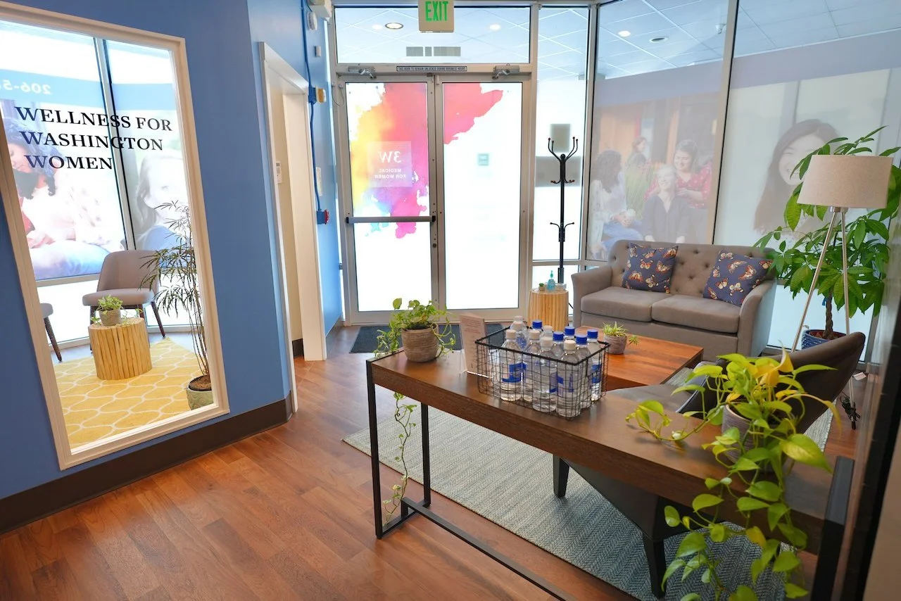 Waiting room with sofa, chairs, plants, and water bottles on a table, near glass doors and windows with group photos and a sign that says "Wellness for Washington Women."