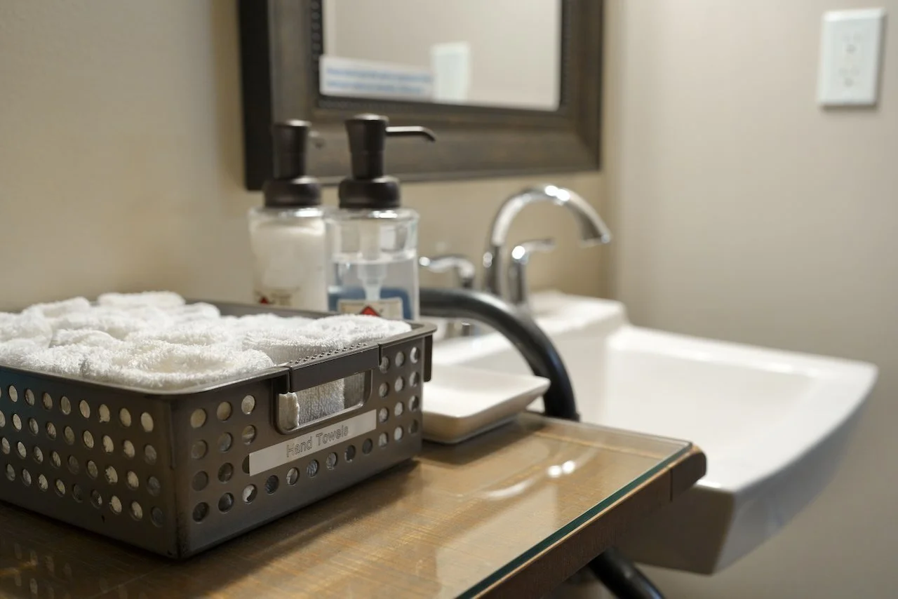 Backsplash of a bathroom with a wooden counter, with a plastic container of white towels, two clear soap dispensers, and a mirror above a white ceramic sink with a chrome faucet.