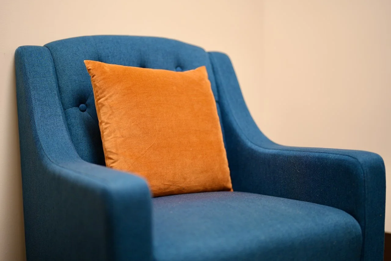 Blue upholstered armchair with an orange pillow on it, against a plain beige wall.