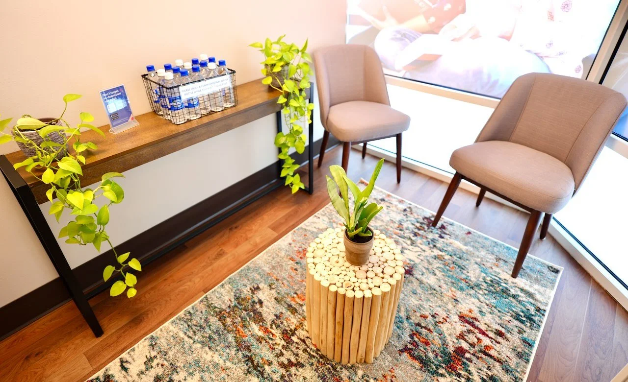 A waiting area with two beige chairs, a potted plant on a wooden table, and another plant on the floor. There is a sideboard with bottled water and informational materials, and large windows allowing natural light.
