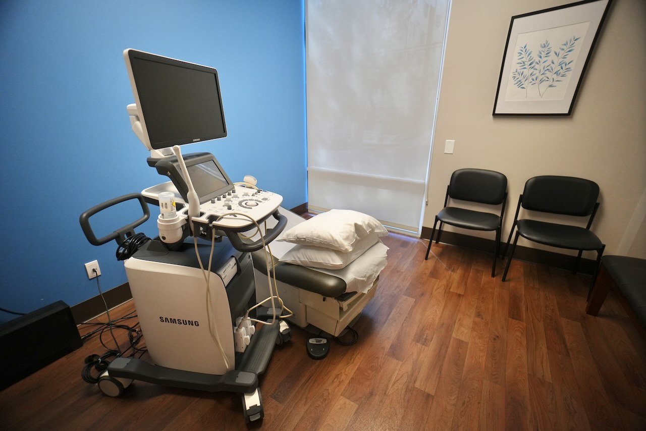 Medical examination room with ultrasound machine, pillows on examination table, and three black chairs against the wall.