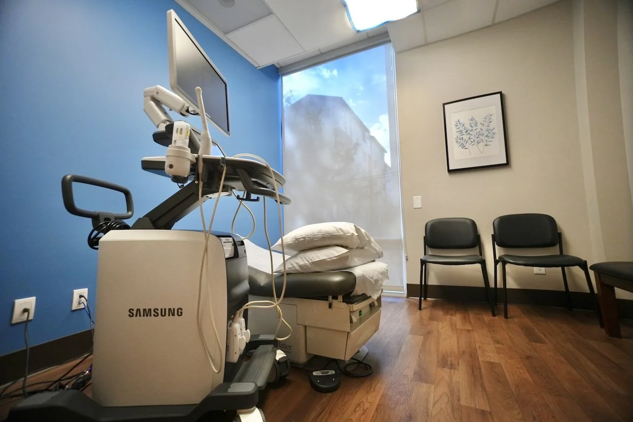 Hospital examination room with a Samsung ultrasound machine, a hospital bed with pillows, and three black chairs against the wall. There is a large window with a sheer curtain showing the outside.
