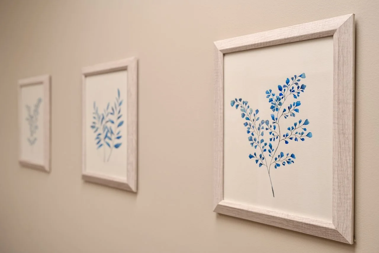 Three framed watercolor paintings of blue leafy branches hanging on a beige wall, viewed from an angle.