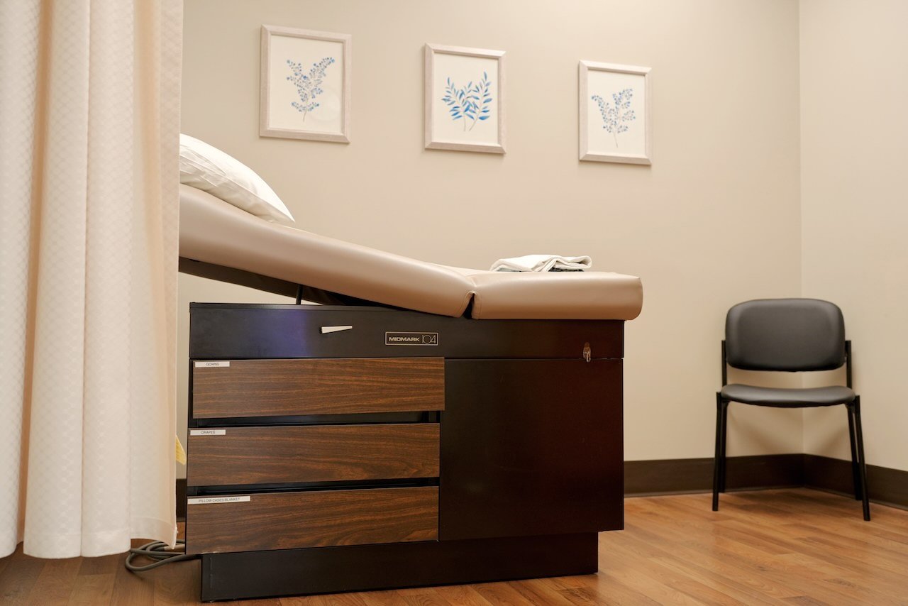 Empty hospital examination room with a bed, a black wooden storage cabinet, a gray chair, and three framed botanical prints on the wall.