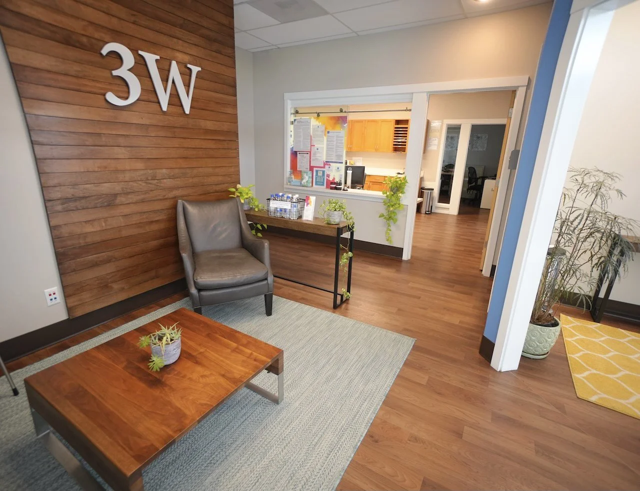 Office waiting area with a wooden accent wall featuring large white letters '3W', a gray armchair, a wooden coffee table with a small potted plant, a side table with bottled water and plants, and a view into a kitchen and other office rooms with hard